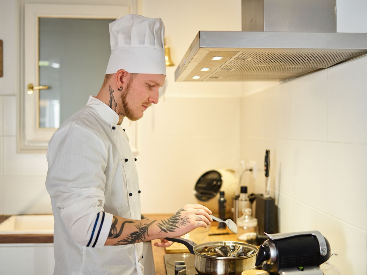 A tattooed chef wearing a white uniform and hat cooking in a contemporary kitchen.